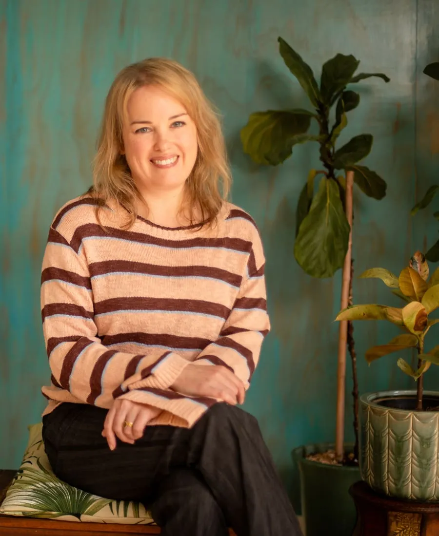 Jo a blonde white woman in her 40s is wearing a stripy jumper with a blue wall and some plants in the background. She is smiling.