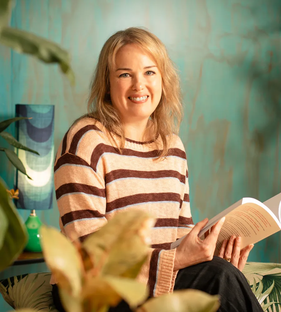 Jo a blonde white woman in her 40s is wearing a stripy jumper with a blue wall and plants in the background. She is smiling and reading her book eight limbs of aerial yoga.