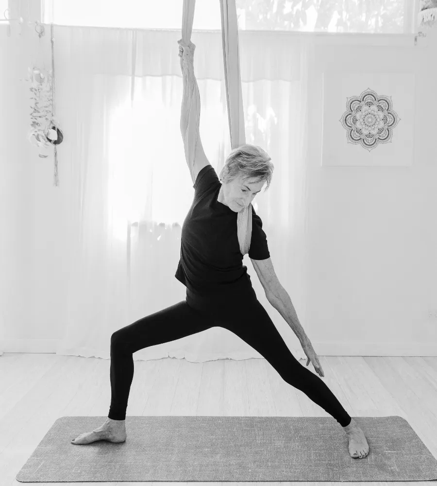 A black and white photograph of Maggie practicing a warrior pose with the aerial hammock wrapped around her back and shoulders.