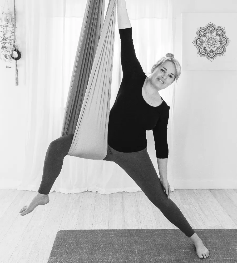 A black and white photograph of Jo, a blonde woman in her 40s doing an aerial yoga warrior pose