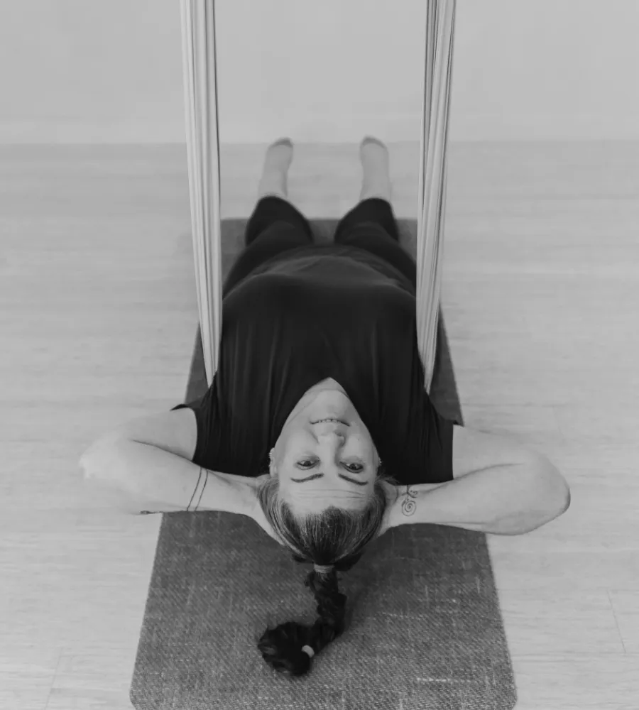 A black and white photograph of Wendy sitting on the ground and leaning back into an aerial hammock to look at the camera.